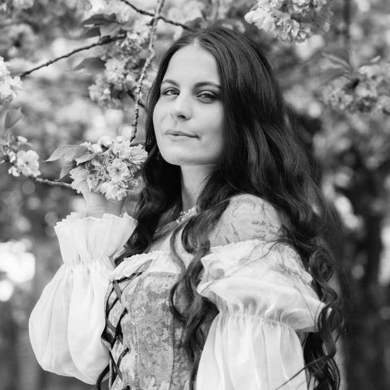 Portraitfotografie Eine junge Frau mit offenen langen, welligen und dunklen Haaren lächelt in die Kamera während sie Kirschblüten festhält.